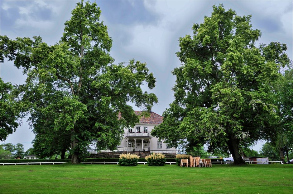 A large white detached house with multiple windows and a prominent gabled roof is shown in the image, set against a background of leafless trees under a partly cloudy sky. The house features a porch a