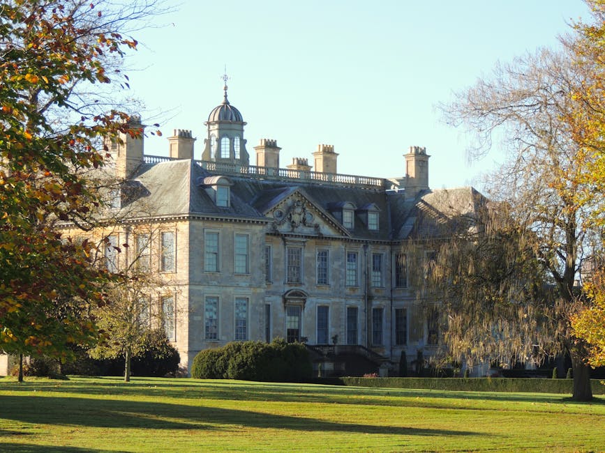A four-storey white residential building with multiple symmetrical windows and a central entrance, situated behind a landscaped lawn with a paved pathway. In the foreground, tree branches and leaves p