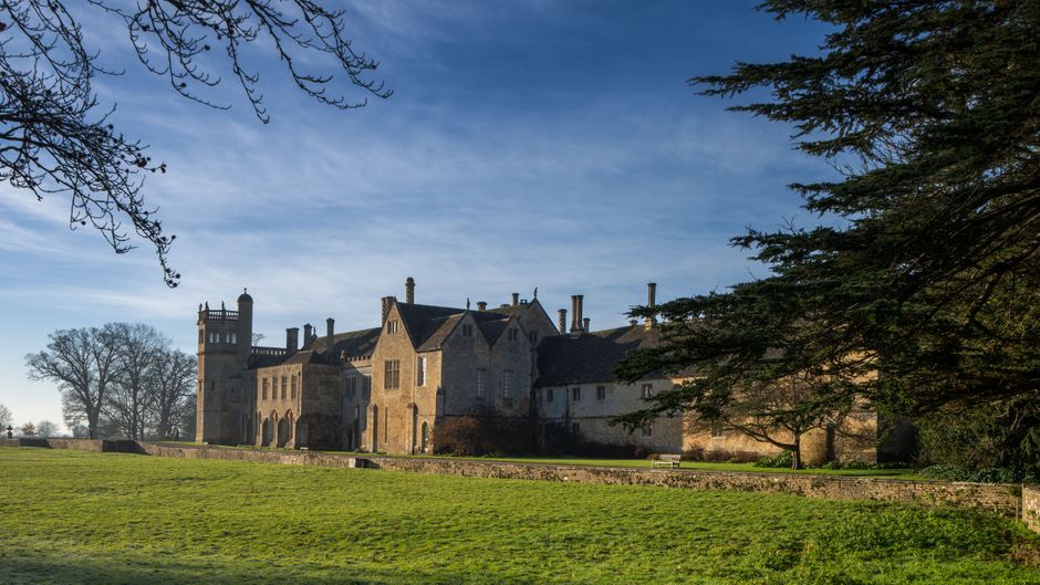 A large historic stone castle with multiple turrets, chimneys, and pitched roofs is visible in the image, set against a bright blue sky with some wispy clouds. The castle is surrounded by a well-maint