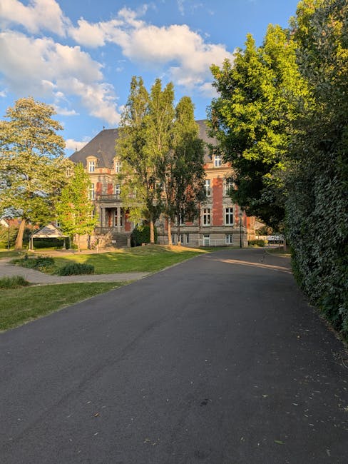 A wide view of a paved driveway leading to a large historic brick building with multiple floors, decorative stone window frames, and a dark slate roof, situated within a green residential area. The scene includes tall, leafy trees on both sides of the driveway, providing shade and framing the building, with some trees displaying fresh, green foliage. To the left, there is a patch of grass with a small pathway, while on the right, a dense hedge runs parallel to the driveway. The sky above is mostly blue with scattered white clouds, and the lighting suggests daytime with clear weather. This setting resembles a peaceful neighborhood or estate where house removals or relocations might take place, and the scene subtly supports the concept of home moving logistics, such as a property prepared for a furniture transport or loading process by a professional removal service like Man and Van Manor House.