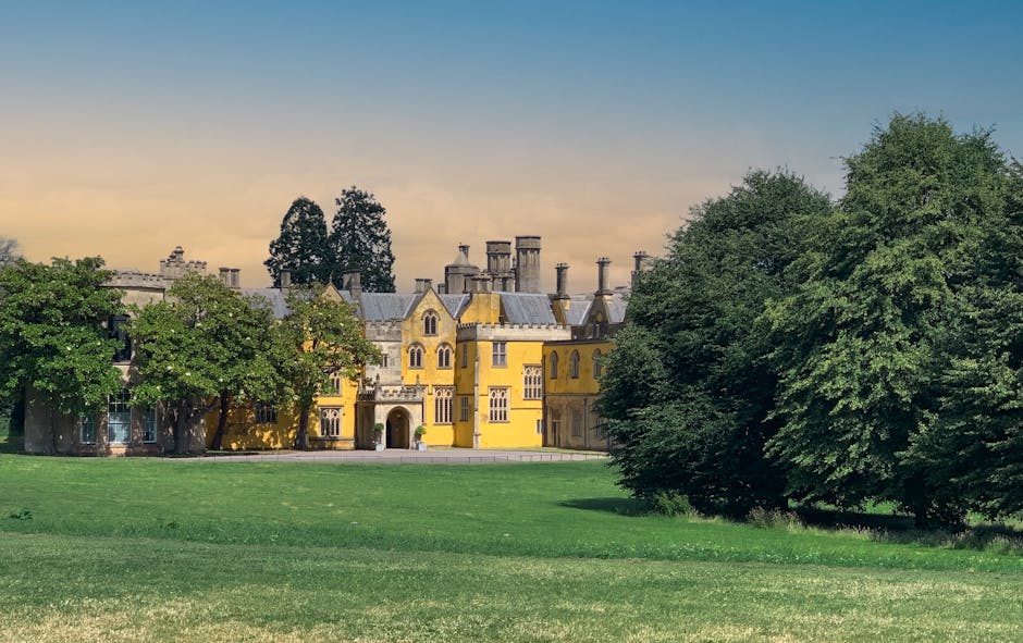 A large, historic yellow mansion with multiple chimneys and ornate architectural details, situated behind a well-maintained green lawn and bordered by tall, leafy trees. The scene is captured during daylight with soft, natural lighting highlighting the building’s features. The mansion appears to be part of a property involved in house removals or relocation services, as there are no visible moving vehicles or equipment in the immediate foreground. The image's setting suggests an environment suitable for furniture transport and packing during a home relocation process, with the house's exterior ready for potential loading and transportation tasks managed by Man and Van Manor House, specialists in removals within the local area.