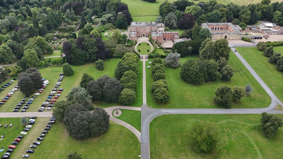 A large, historic yellow castle with multiple turrets and stone chimneys, situated behind a extensive green lawn and flanked by two large leafy trees. The scene is captured during daylight with a part