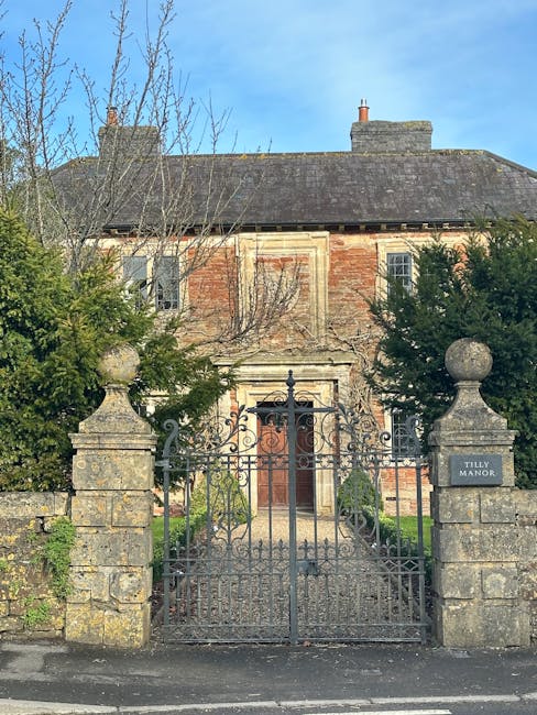 A large Victorian-style brick house with multiple steeply pitched roofs, turrets, and tall brick chimneys, situated behind a stone wall and white gate on a residential street. The property features or