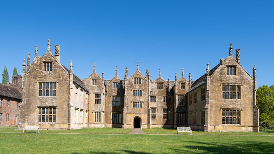A large historic stone building with multiple gabled roofs, tall mullioned windows, and decorative chimneys, situated behind a well-maintained grassy lawn with two white benches. The building is part of a property requiring house removals and relocation services, with an entrance visible in the centre. The scene is set outdoors on a clear day with a bright blue sky and surrounding green trees, indicating a peaceful setting suitable for home relocation. The image reflects the type of property often involved in furniture transport and packing and moving activities, which Man and Van Manor House provides, showcasing the exterior environment where loading and unloading could occur during a move.